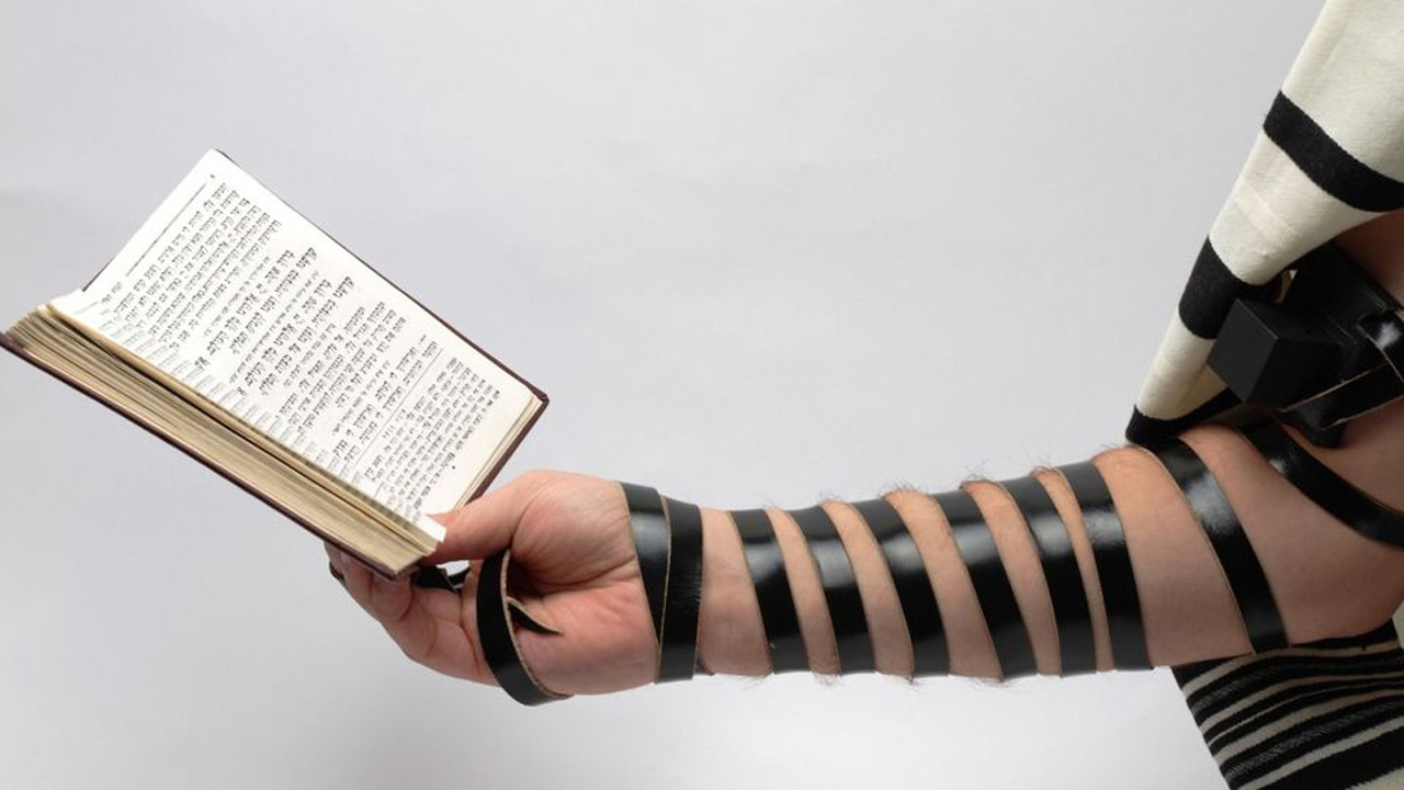 An image of an arm wrapped in tefillin holding a prayer book.